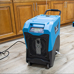A blue and black portable dehumidifier unit on a light wood floor in front of wooden kitchen cabinets, with a power cord and a second blue device in the background.